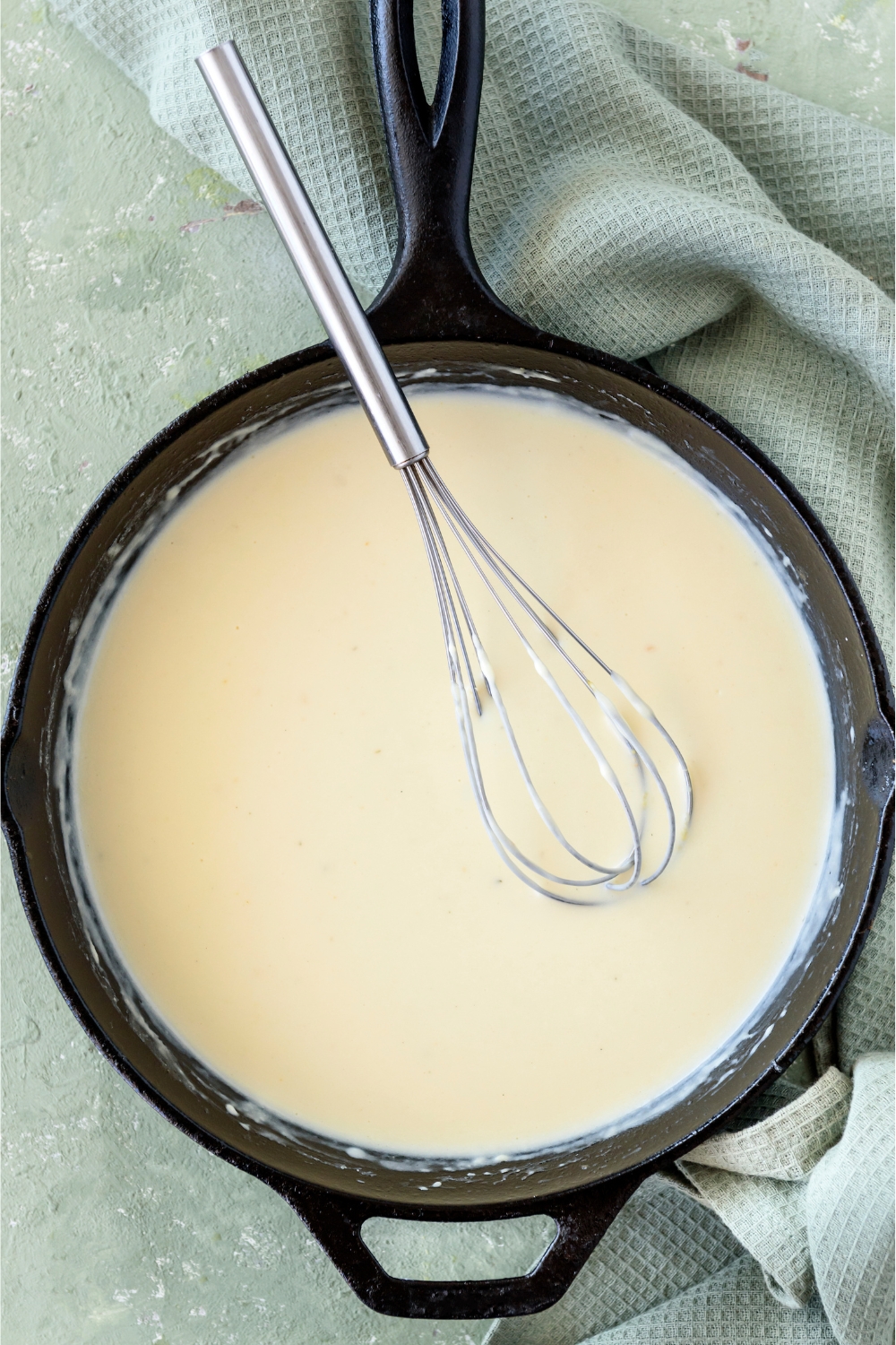 A cast iron skillet filled with cream sauce and a whisk is in the skillet.