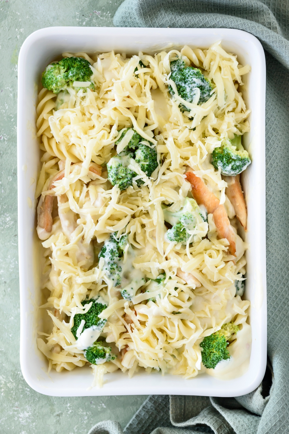 A baking dish filled with cooked pasta, broccoli, and chicken strips in a creamy sauce topped with shredded cheese.