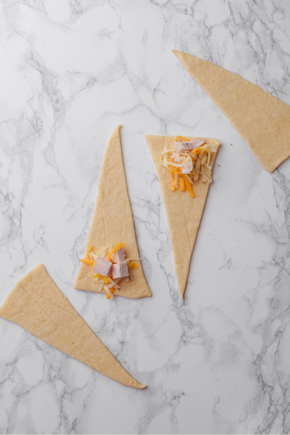 Four crescent dough triangles on a grey counter and two have diced chicken and shredded cheese on them.