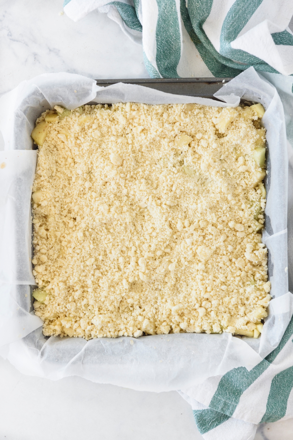 A square baking dish lined with parchment paper filled with unbaked apple pie bars and a crumble topping.