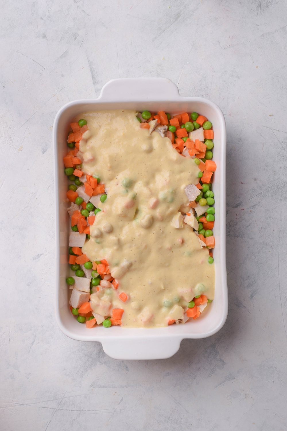 A baking dish filled with diced chicken, peas, and carrots, and a creamy soup mixture has been poured on top.