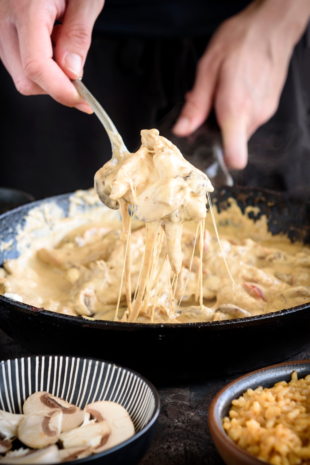 A spoonful of creamy chicken and vegetables with cheese stringing from the spoon to the skillet below.