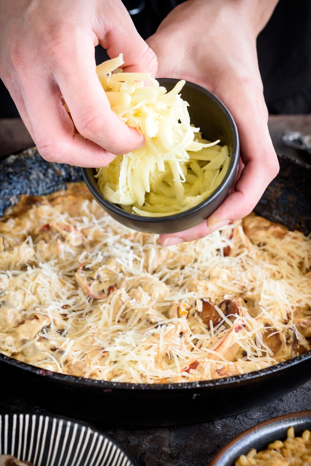 A person holding a bowl of cheese and sprinkling it over a skillet filled with chicken, peppers, and mushrooms in a cream sauce.