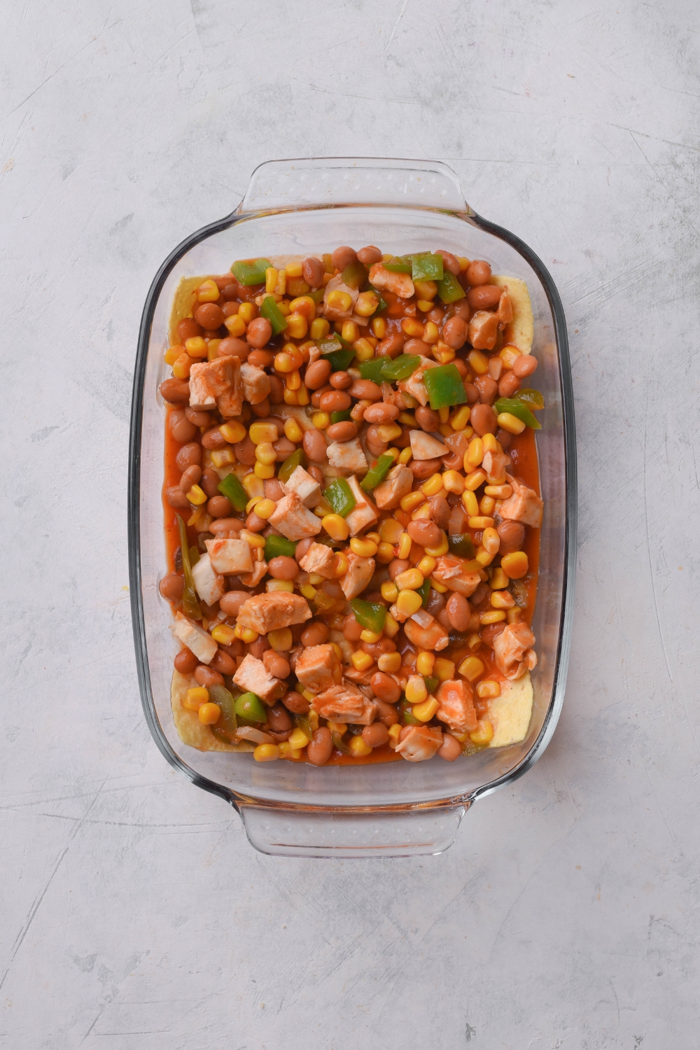 A baking dish filled with pinto beans, diced green peppers, diced onion, corn, and diced chicken in sauce.
