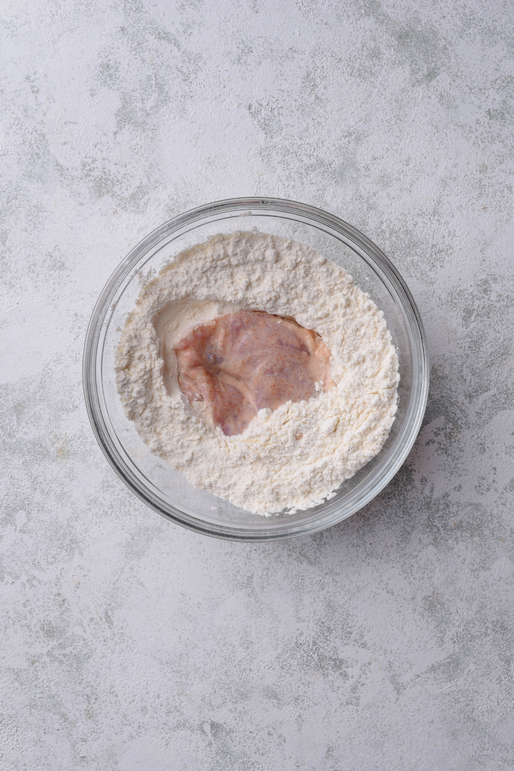 A bowl filled with a flour mixture and a piece of raw chicken.