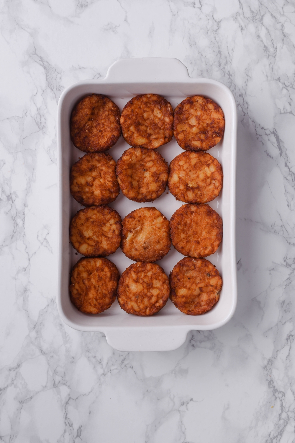 A white baking dish filled with twelve hash brown patties lining the bottom of the dish.