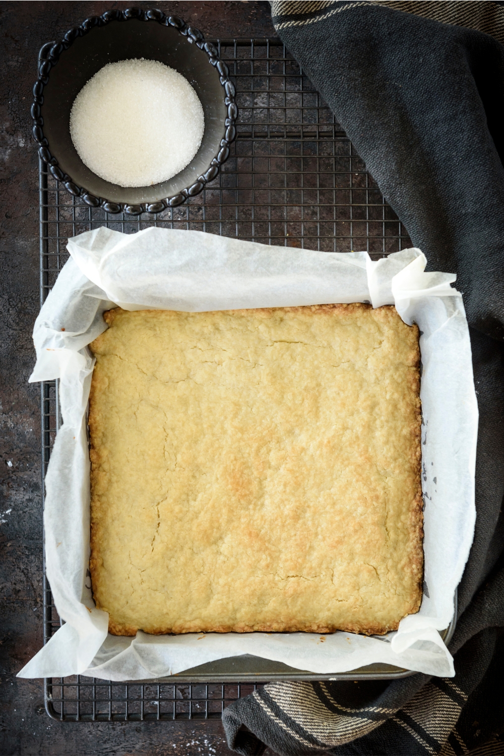 A square baking dish lined with parchment paper and filled with a baked shortbread biscuit on a wire rack next to a bowl of sugar.