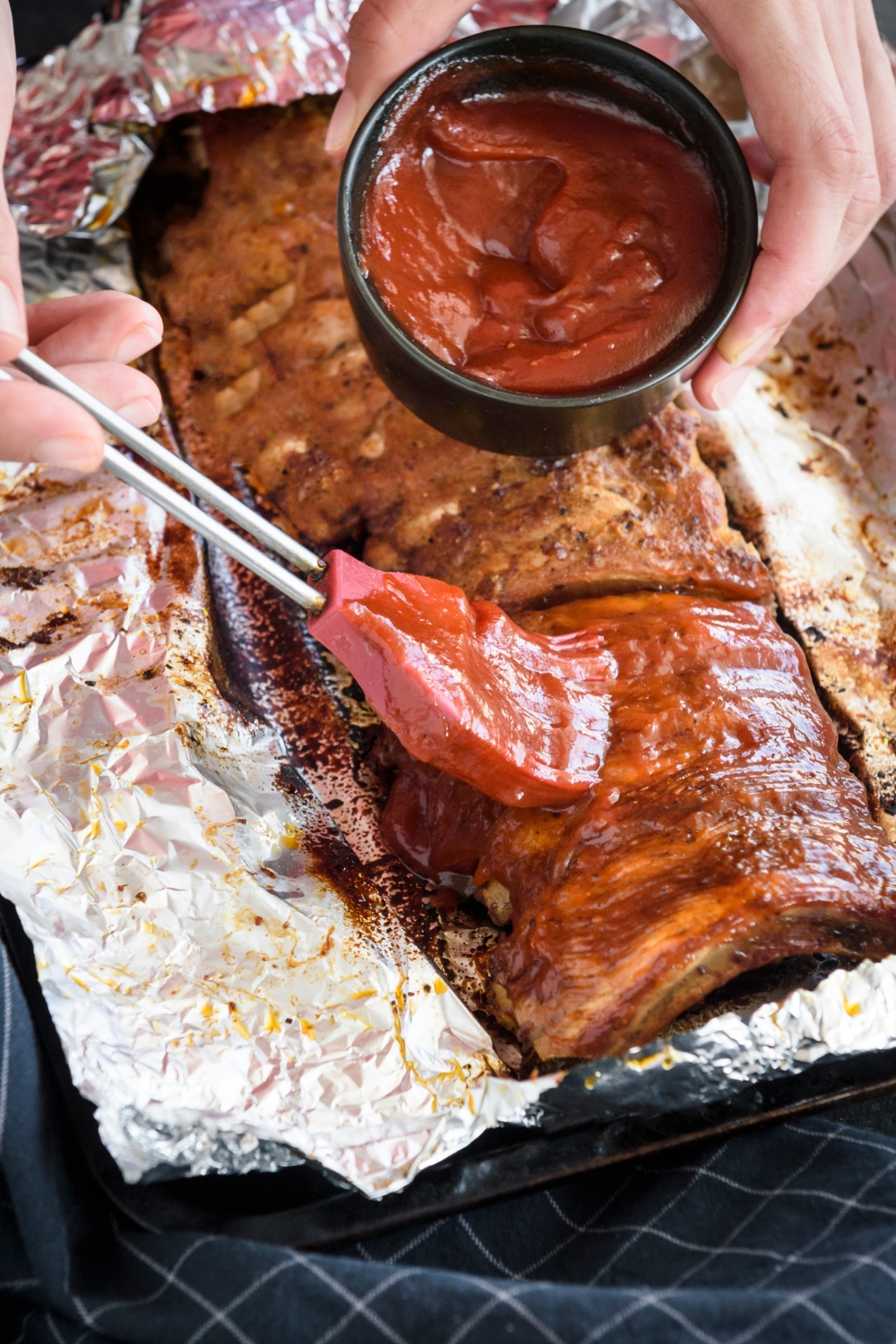 A rack of ribs wrapped in foil being brushed with barbecue sauce.