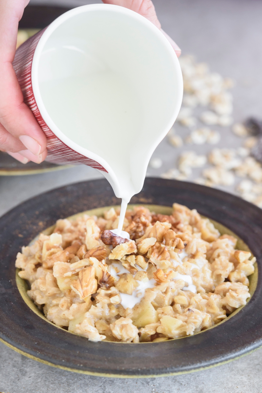 A pitcher of milk being poured over a bowl of oatmeal that's topped with apples and chopped nuts.