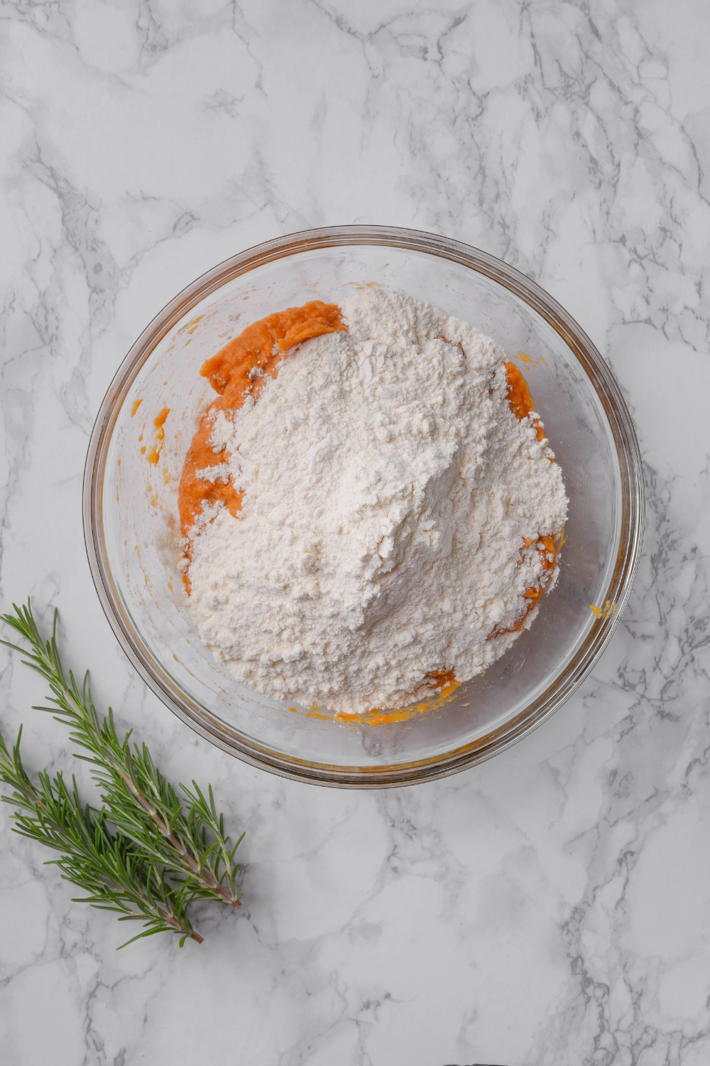 A clear bowl filled with mashed sweet potato and flour. The bowl is on a grey counter next to two sprigs of rosemary.