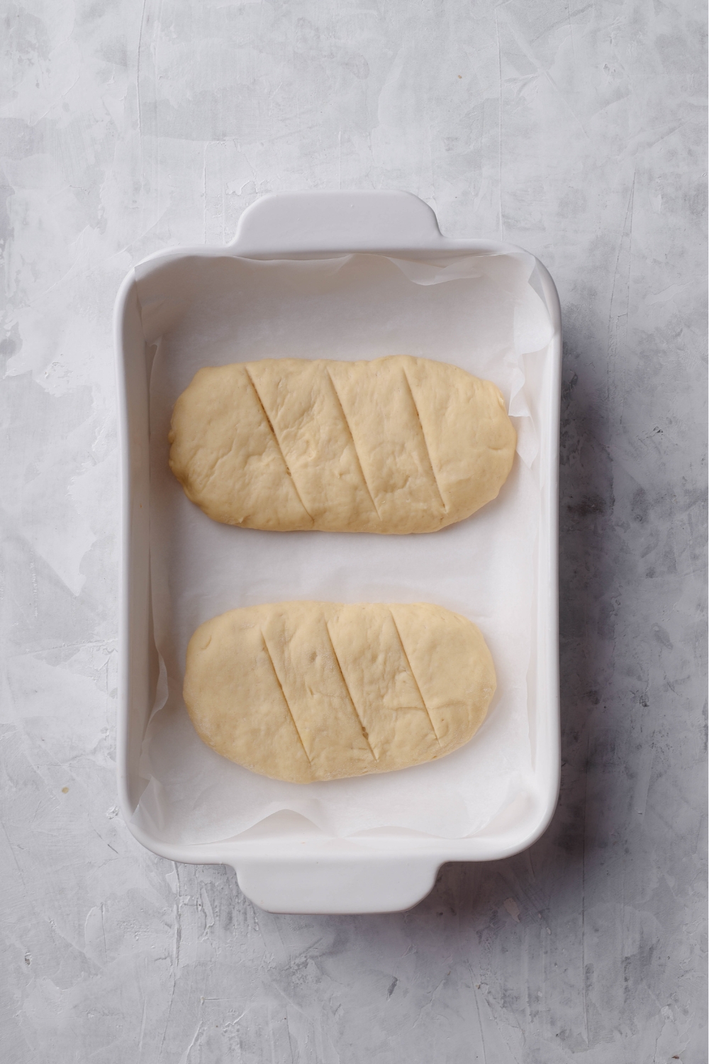 two raw loaves of dough with scores in them in a white baking dish