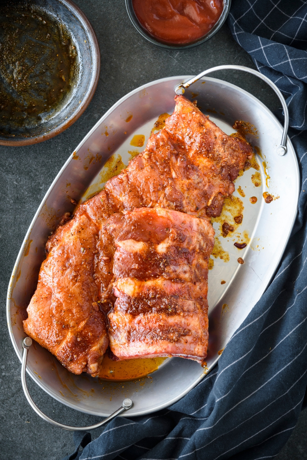 Raw spare ribs covered in oil and a dry seasoning rub in a metal dish.