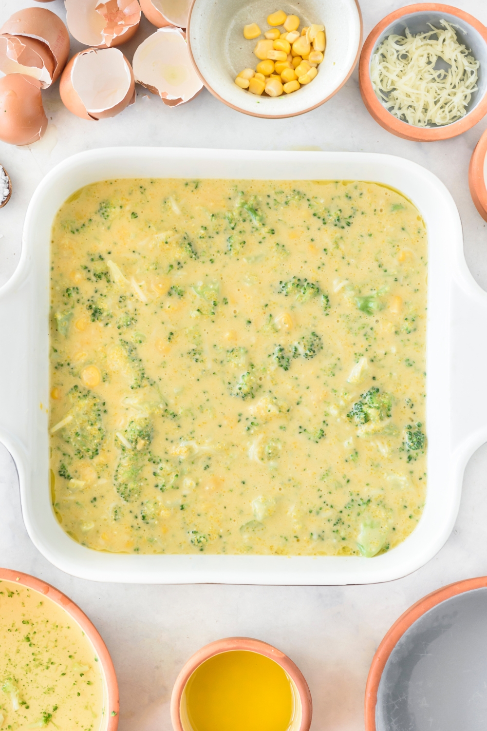 A square baking dish filled with cornbread batter with chunks of broccoli and whole corn kernels.