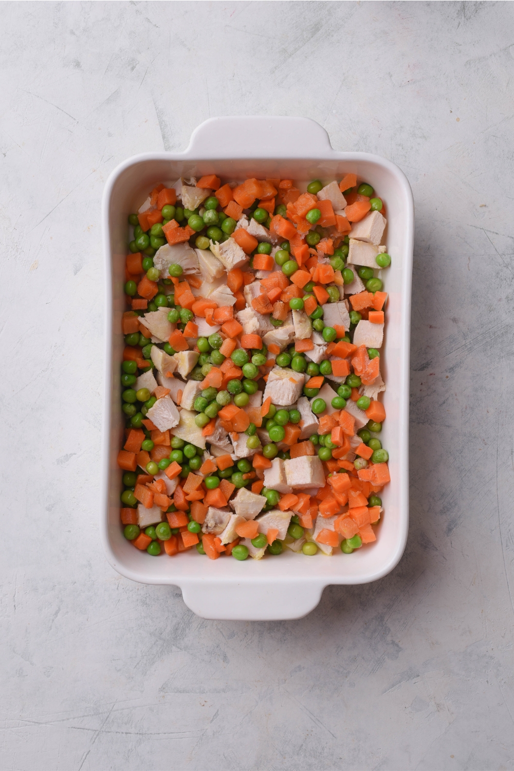 A white baking dish filled with diced chicken, peas, and carrots.