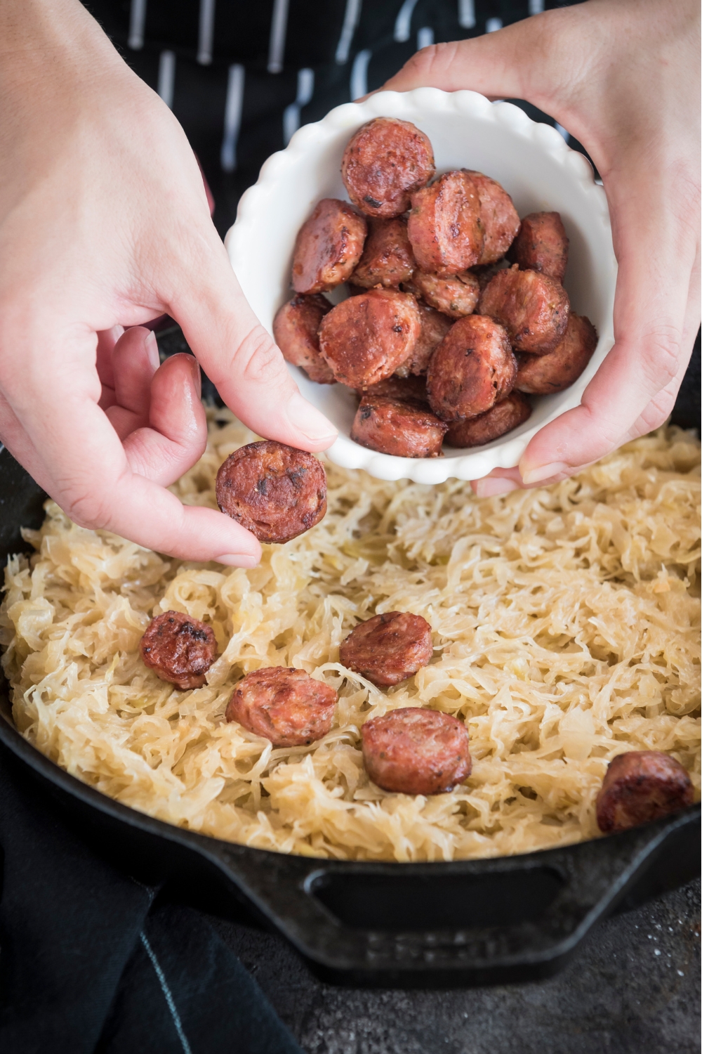 A bowl of sliced sausages being sprinkled over a pan full of sauerkraut.