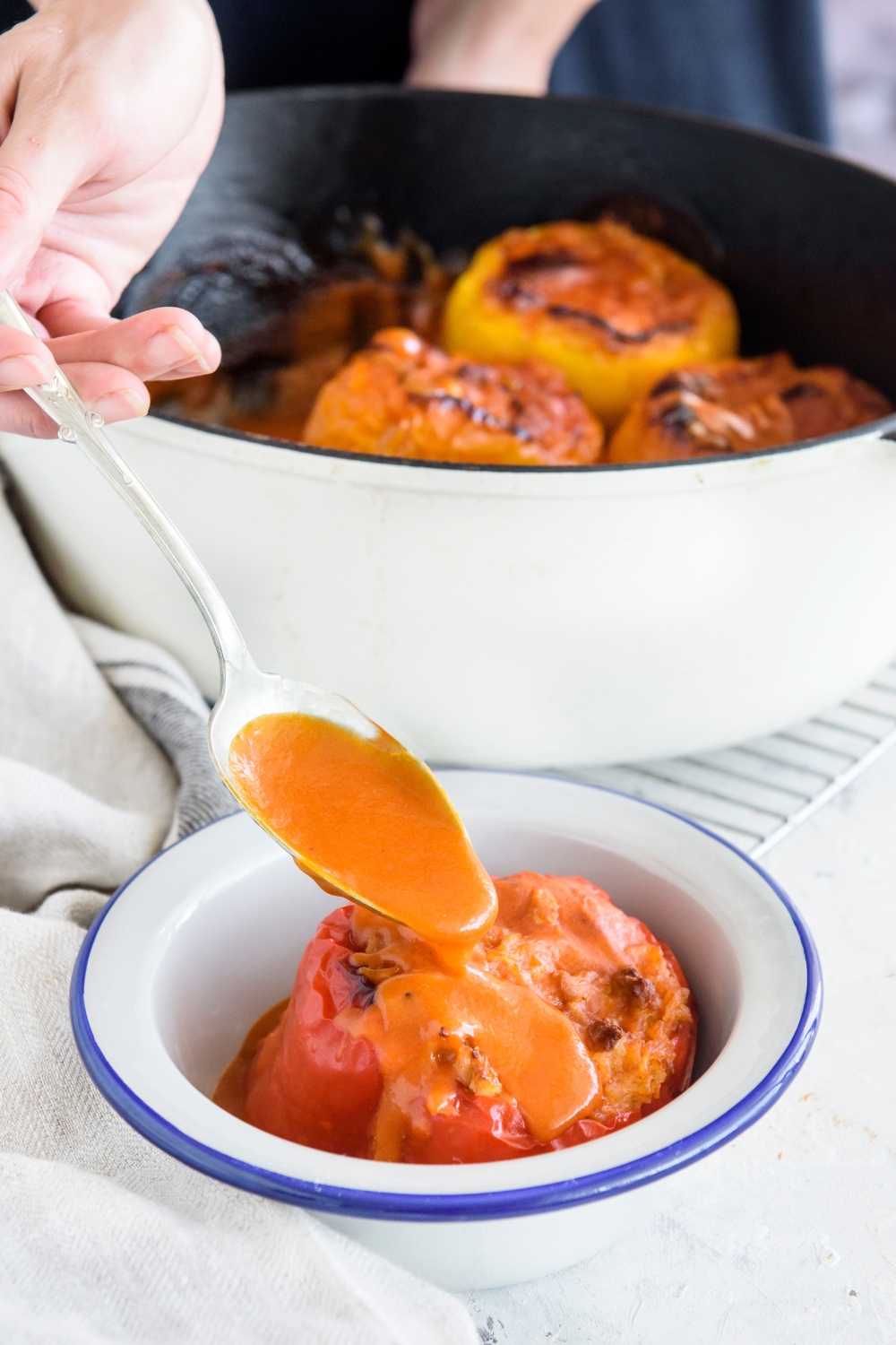 A hand spooning tomato sauce over a stuffed bell pepper. In the background is a baking dish filled with more stuffed peppers.