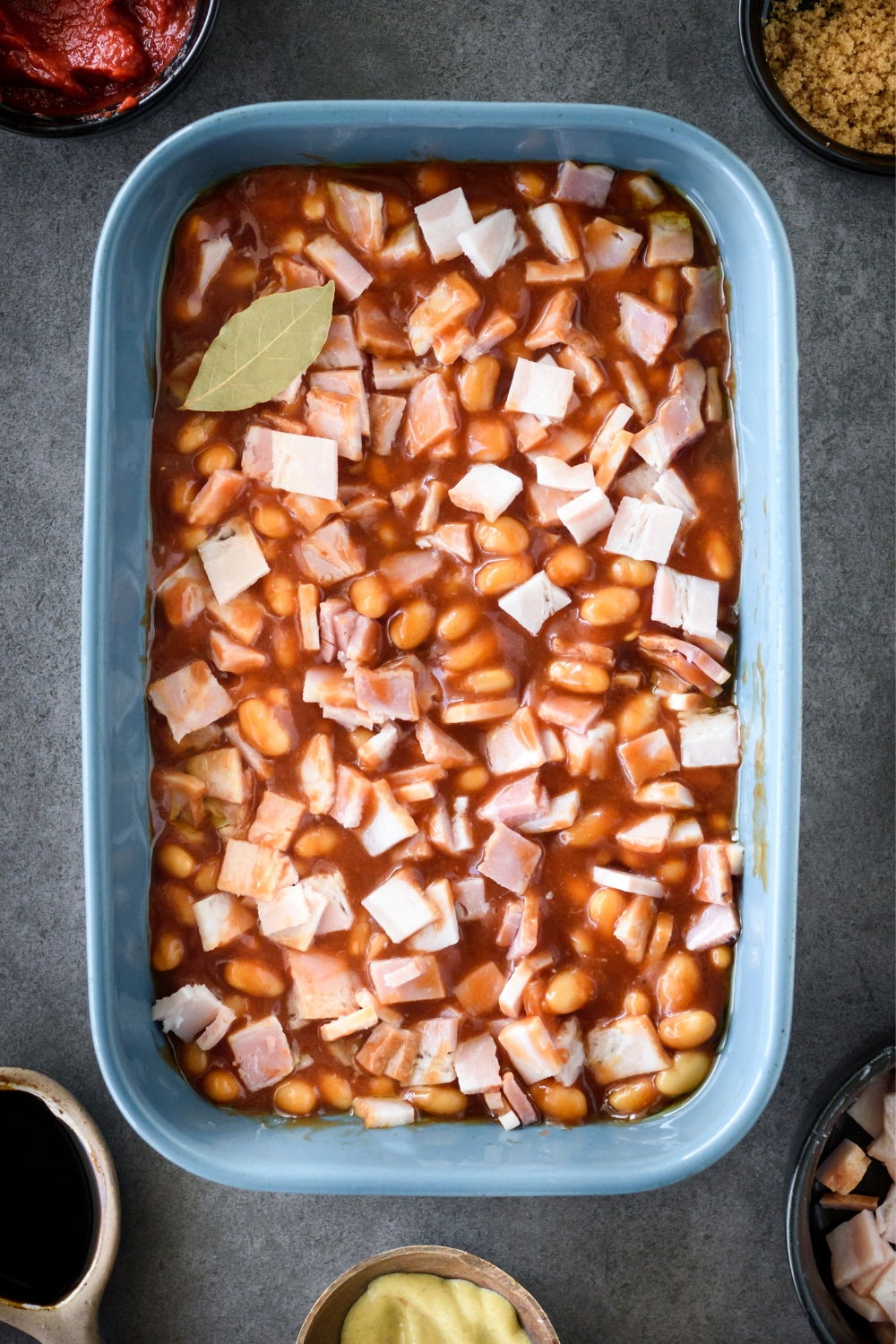A blue casserole dish full of baked beans, bacon, and brown sugar mixture