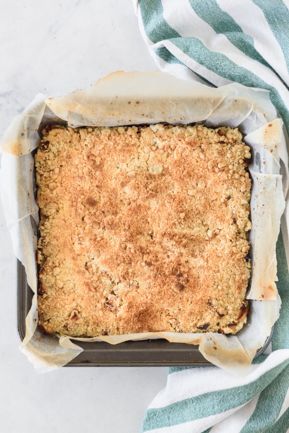 A square baking dish lined with parchment paper filled with freshly baked apple pie bars.