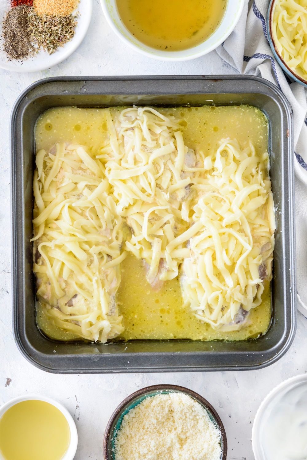 A baking dish filled with three raw chicken breasts topped with shredded cheese and chicken broth is partially covering the chicken.