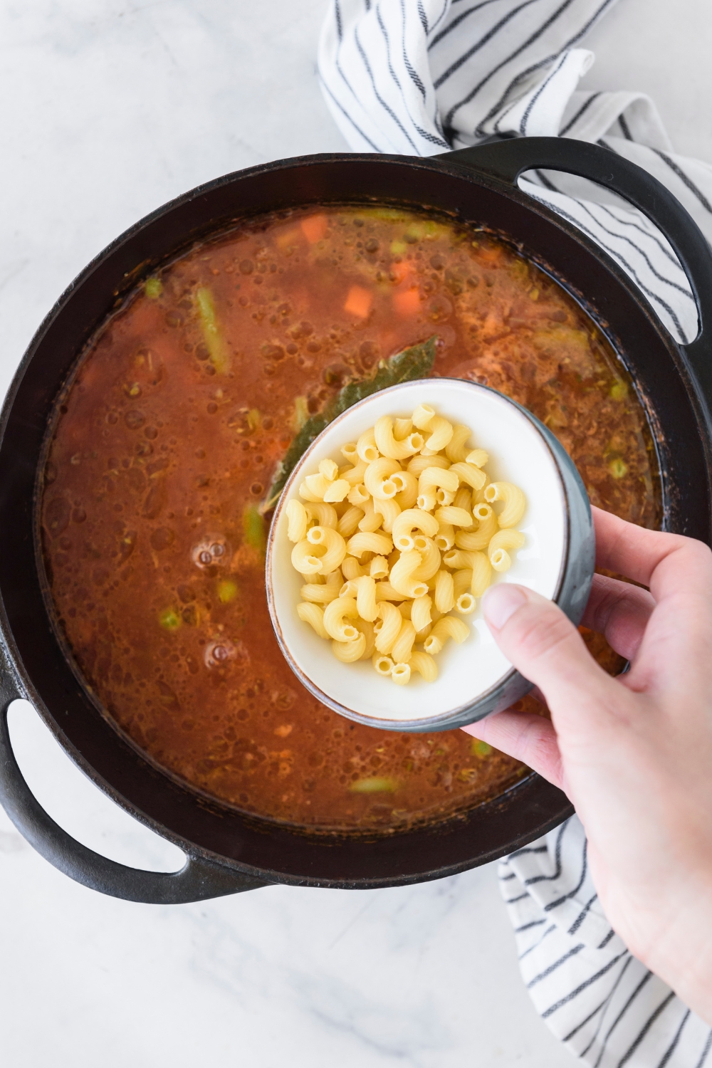 A large pot of broth with mixed vegetables and a bay leaf floating in it and a bowl of dried macaroni noodles is about to be dumped in.