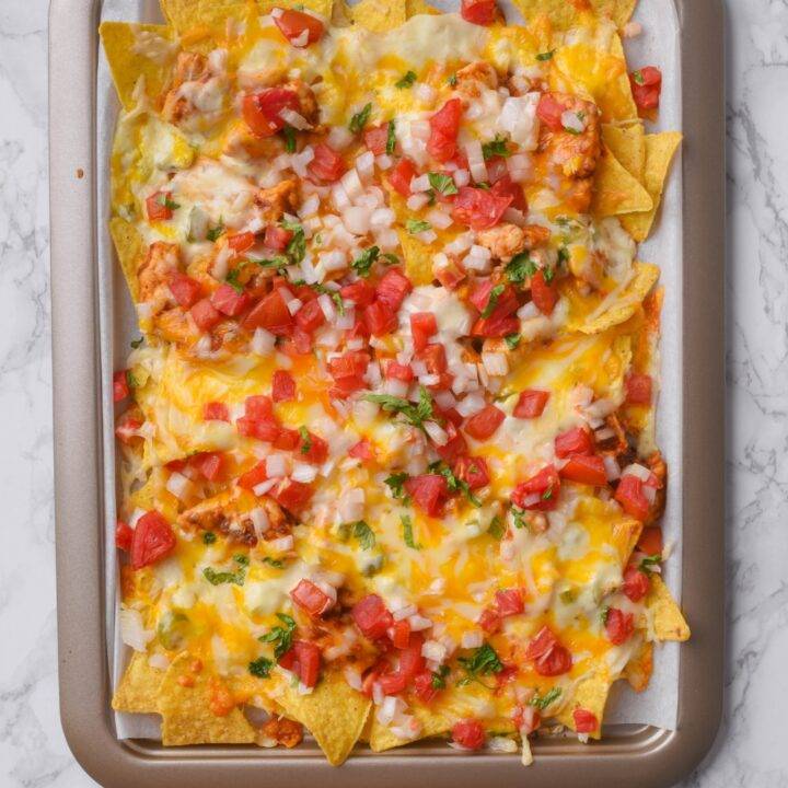 A baking sheet lined with parchment paper topped with tortilla chips, melted cheese, seasoned chicken, and pico de gallo.
