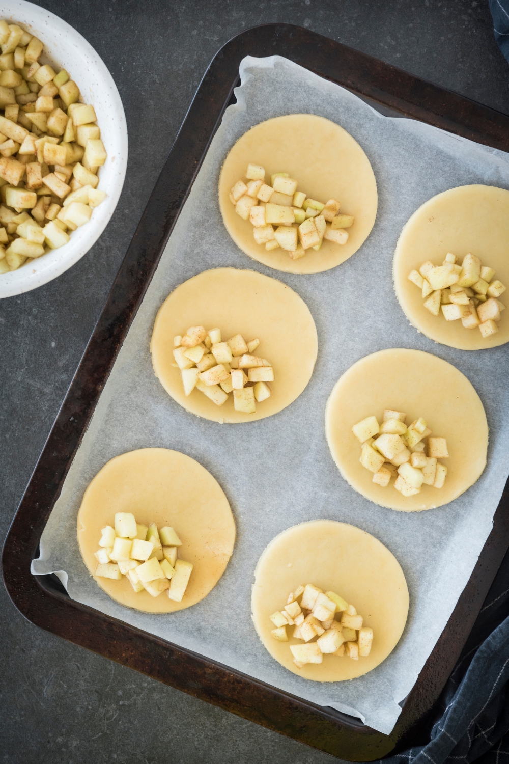 A baking sheet lined with parchment paper with six dough circles each topped with diced apples on one half of the circle.