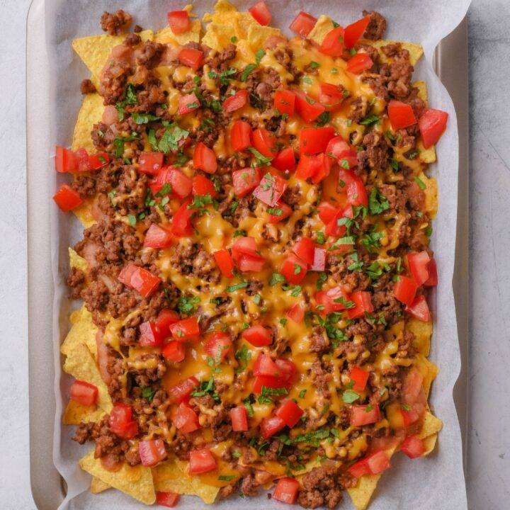 A baking sheet lined with parchment paper topped with tortilla chips, ground beef, melted cheese, chopped tomatoes, and cilantro.