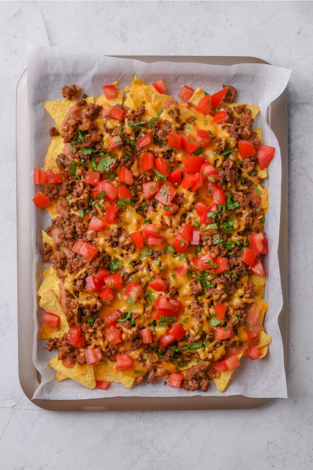 A baking sheet lined with parchment paper topped with tortilla chips, ground beef, melted cheese, chopped tomatoes, and cilantro.