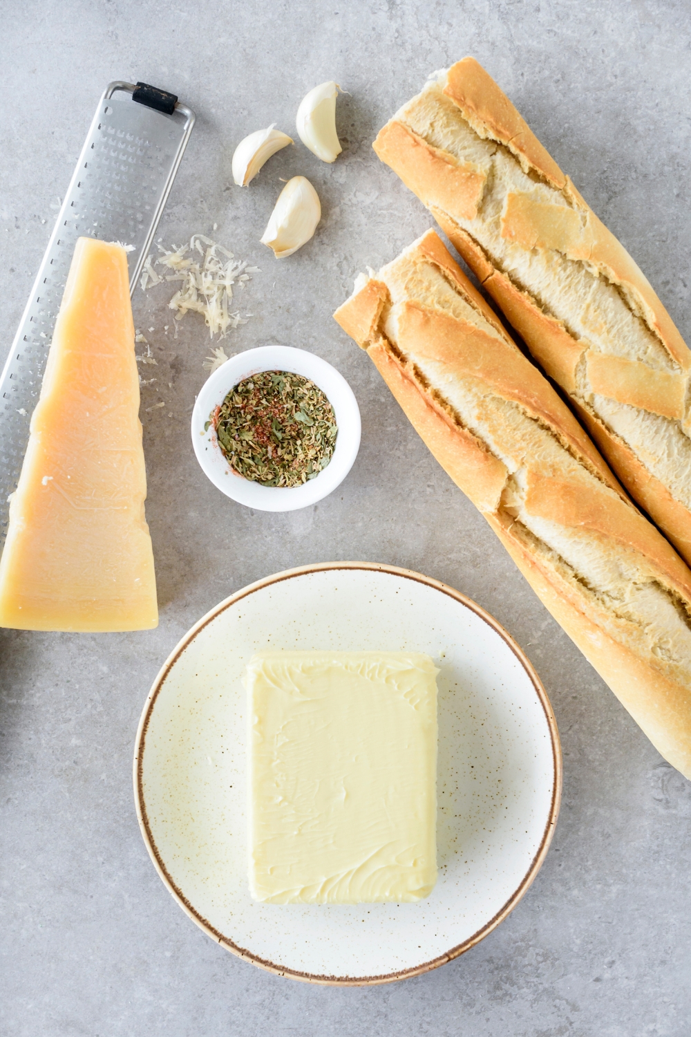 A plate of butter next to a wedge of parmesan cheese, a baguette cut in half, three garlic cloves, and a bowl of seasonings.