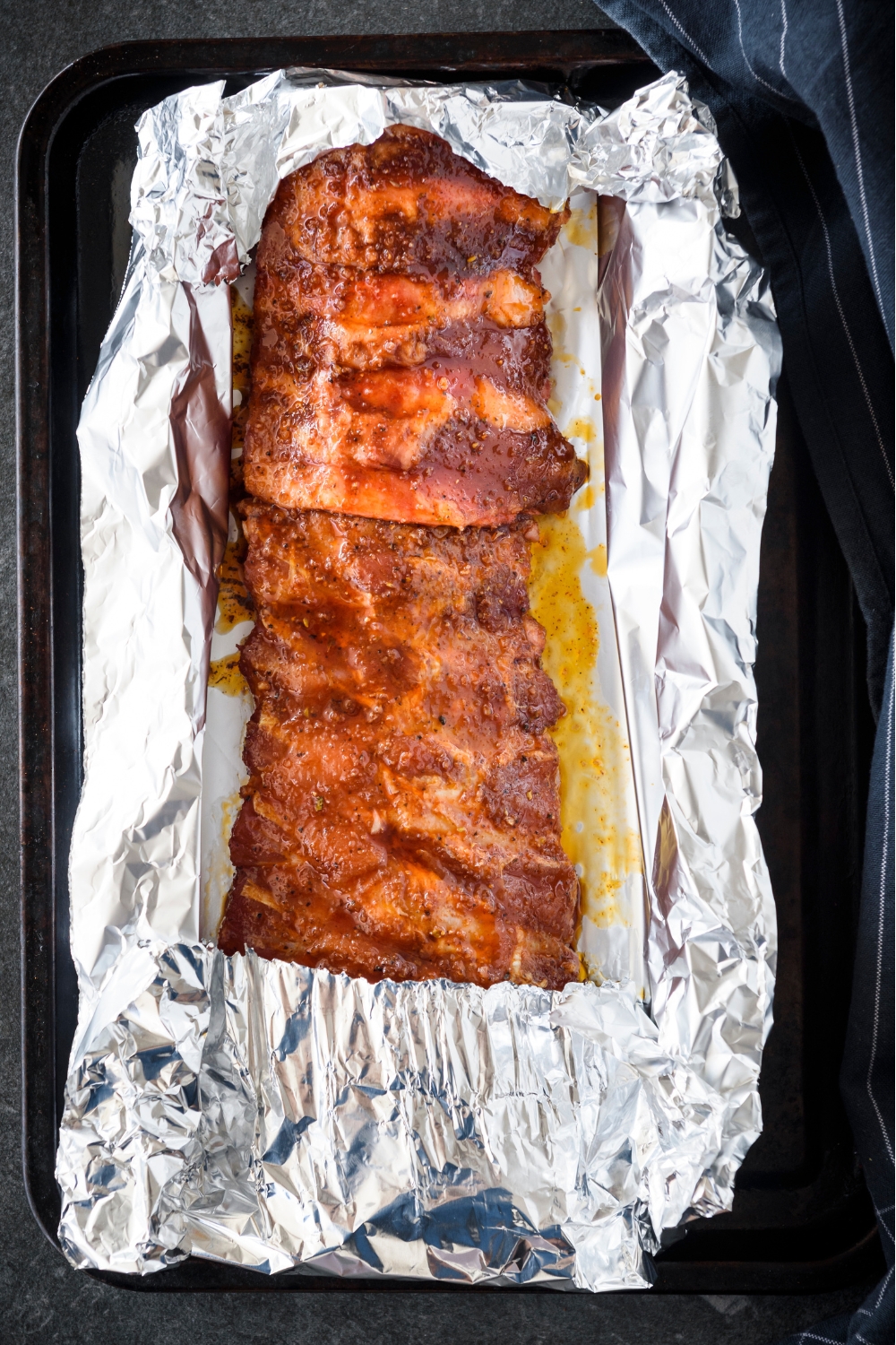 A rack of ribs covered in a seasoning mixture wrapped in foil on a baking sheet.