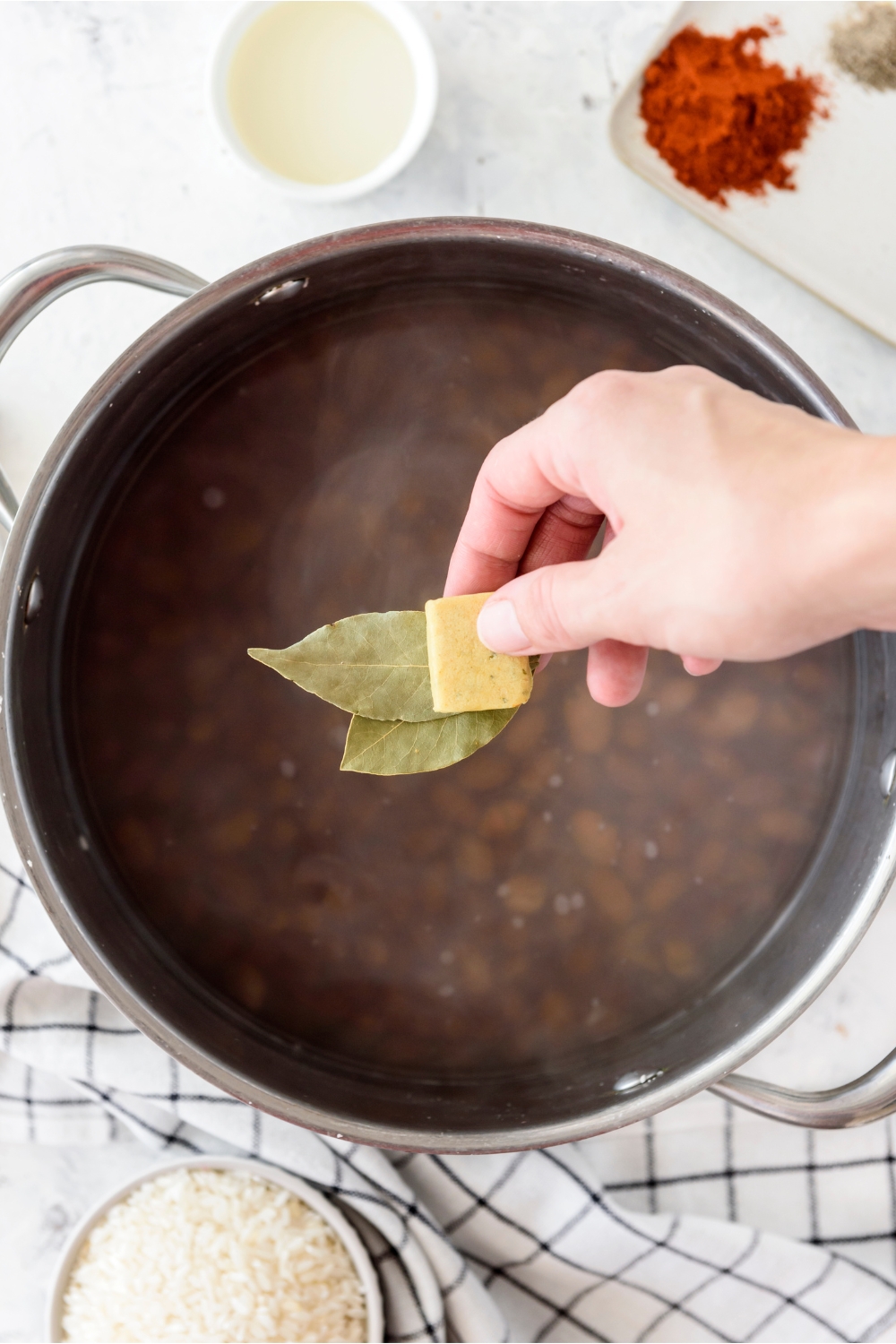 A hand holding a chicken bouillon cube and two bay leaves above a pot filled with beans in water.