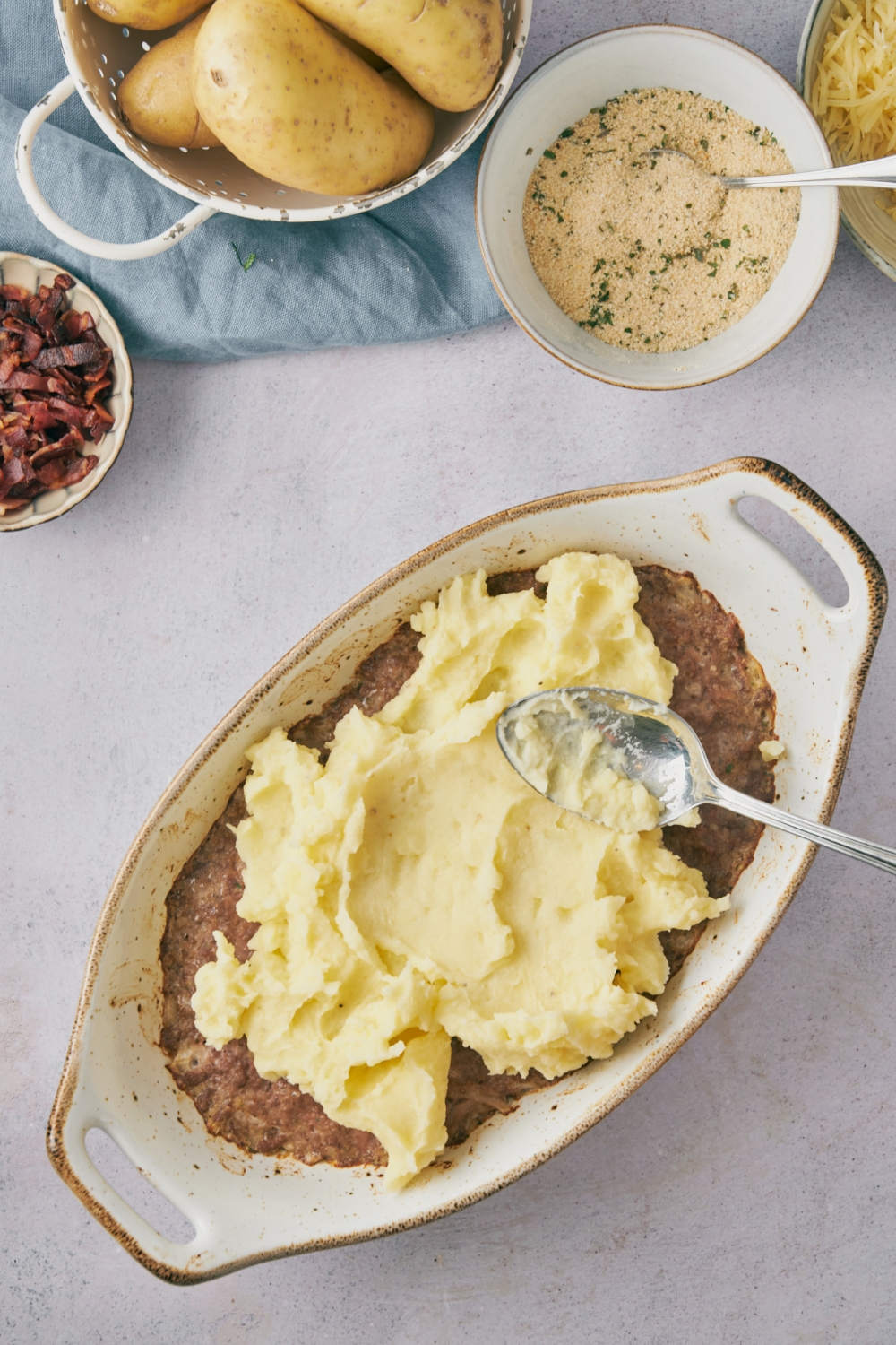 A spoon spreading mashed potatoes over brown meat mixture in a white casserole dish.