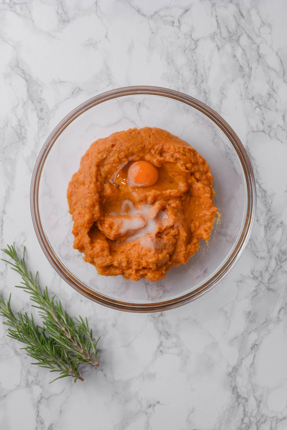 A clear bowl filled with mashed sweet potato and an unbeaten egg. The bowl is on a grey counter next to two sprigs of rosemary.