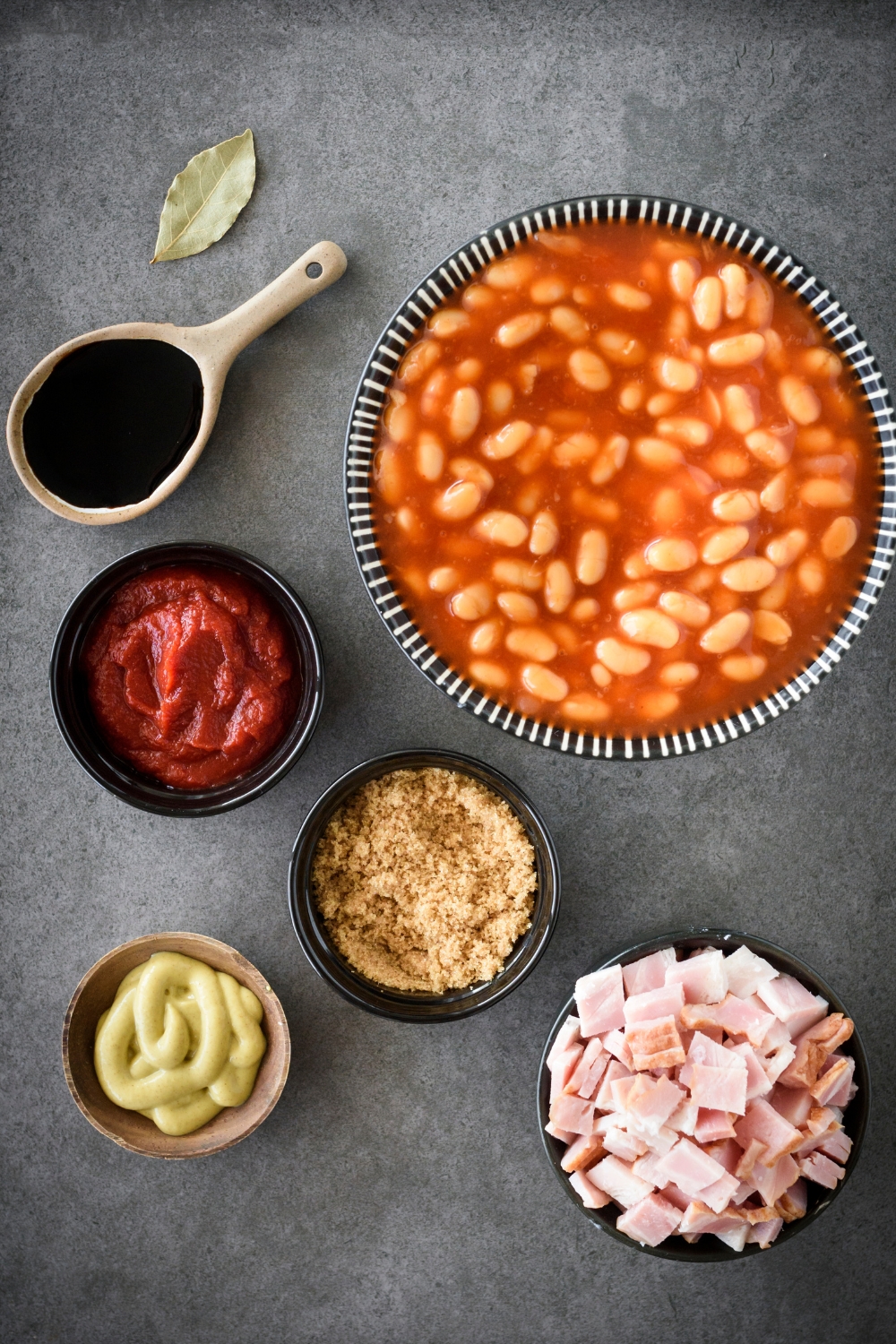 canned baked beans, tomato paste, mustard, diced bacon, and brown sugar in separate bowls on a black counter