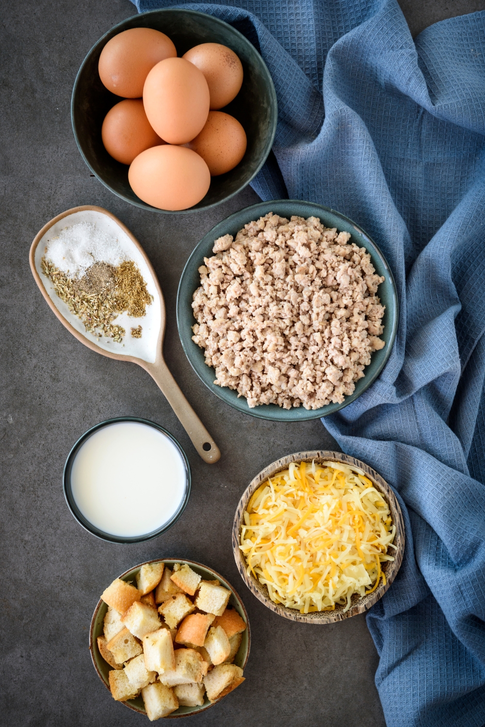 An assortment of ingredients including bowls of eggs, sausage crumbles, shredded cheese, milk, bread cubes, and seasonings.