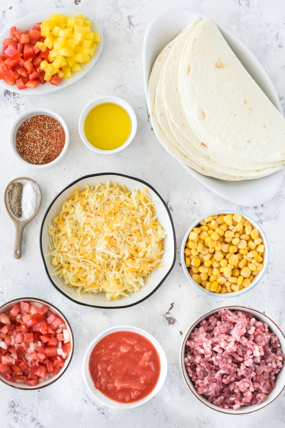 An assortment of ingredients including a plate of tortillas and bowls of shredded cheese, raw ground beef, salsa, corn, diced peppers, oil, and seasonings.