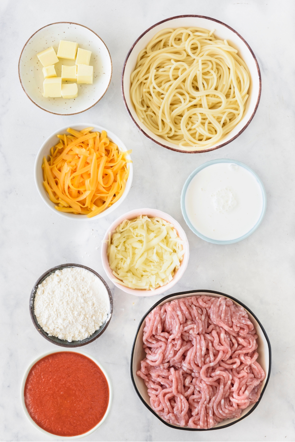 An assortment of ingredients including bowls of cooked spaghetti noodles, tomato sauce, raw ground meat, shredded cheese, flour, cream, and butter.