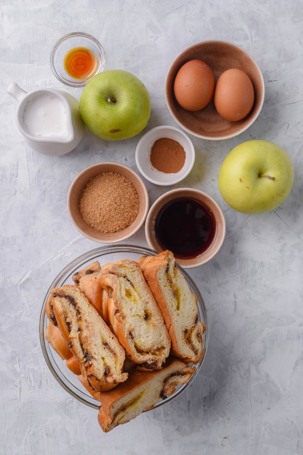 An assortment of ingredients including bowls of bread slices, two eggs, brown sugar, cinnamon, vanilla extract, maple syrup, a pitcher of cream, and two green apples on a grey counter.