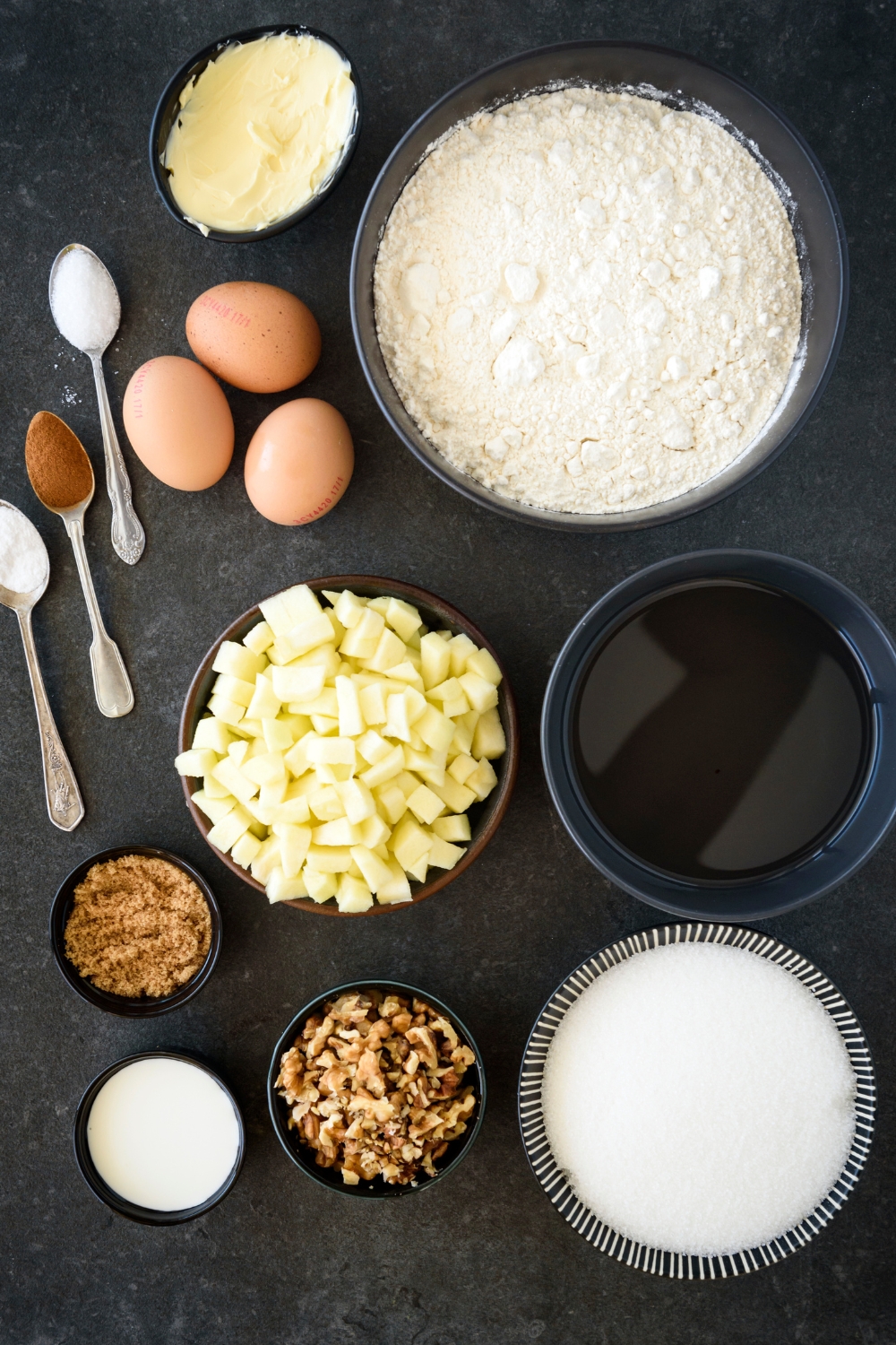 An assortment of ingredients including bowls of flour, butter, diced apples, chopped nuts, sugar, three eggs, and three spoonfuls of spices, all on a black countertop.