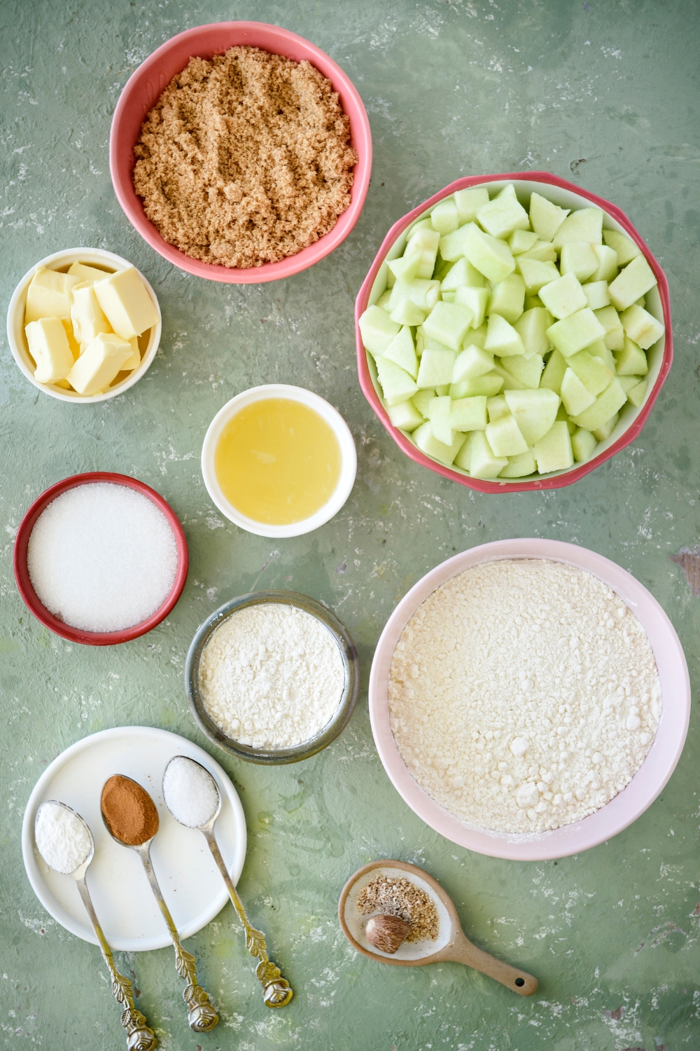 An assortment of ingredients including bowls of flour, diced apples, brown sugar, butter, lemon juice, and spoonfuls of spices resting on a plate. Everything is on a blue countertop.
