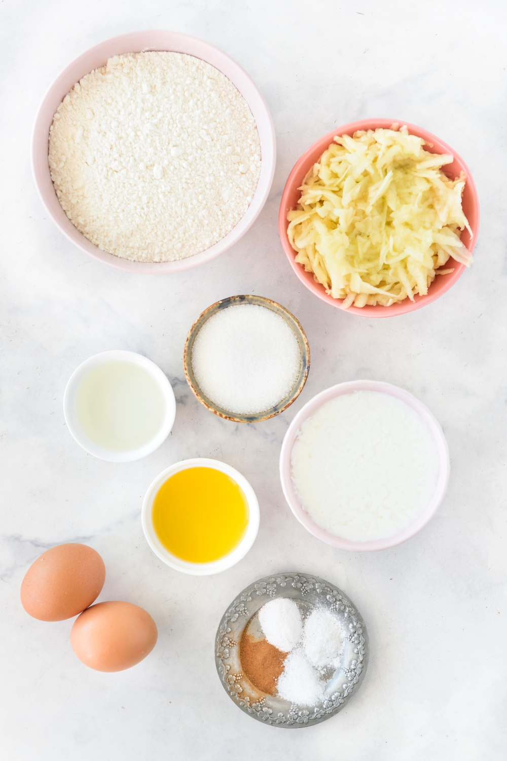 An assortment of ingredients including bowls of flour, grated apple, melted butter, sugar, spices, and two eggs on a white countertop.