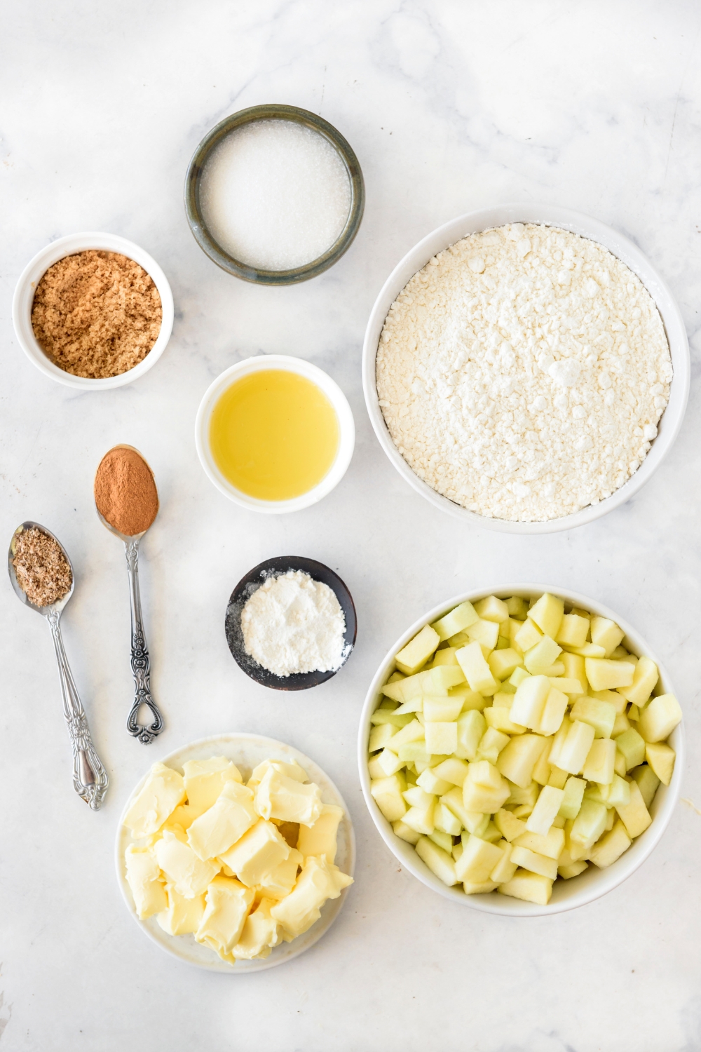 An assortment of ingredients including bowls of diced apples, flour, butter cubes, brown sugar, melted butter, white sugar, and spoonfuls of spices.