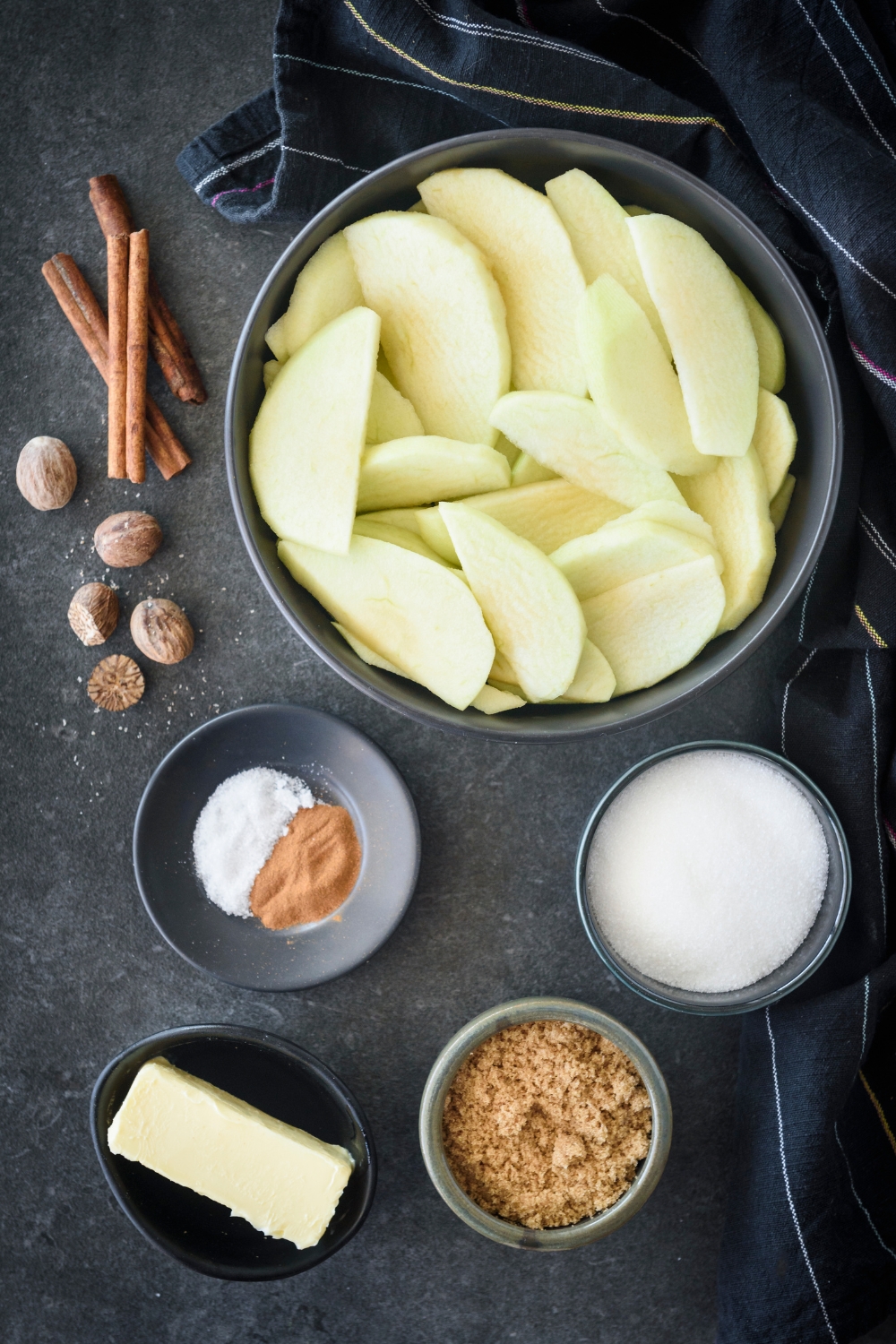 An assortment of ingredients including bowls of apple slices, butter, brown sugar, white sugar, spices, cinnamon sticks, and nutmeg all on a black countertop.