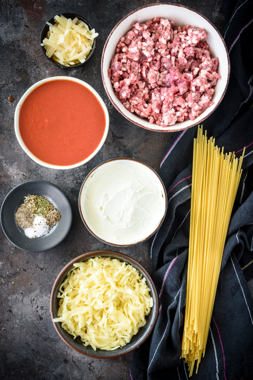 An assortment of ingredients including bowls of raw ground beef, pasta sauce, cream cheese, shredded cheese, spices, and a bundle of dried spaghetti noodles.