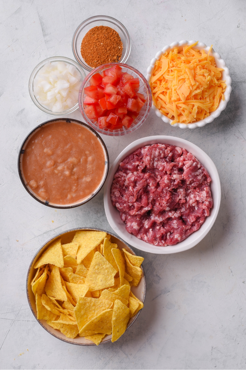 An assortment of ingredients including bowls of raw ground beef, shredded cheese, refried beans, chopped tomatoes, diced onions, and tortilla chips.