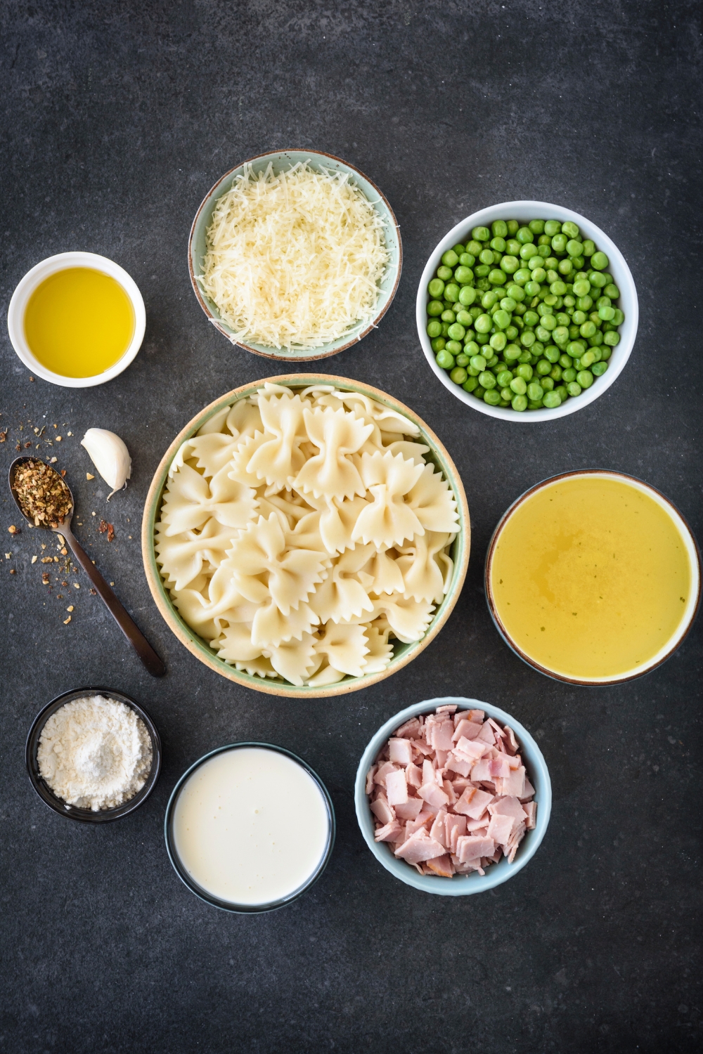 An assortment of ingredients including bowls of bow tie pasta, diced ham, peas, shredded cheese, flour, milk, and spices.