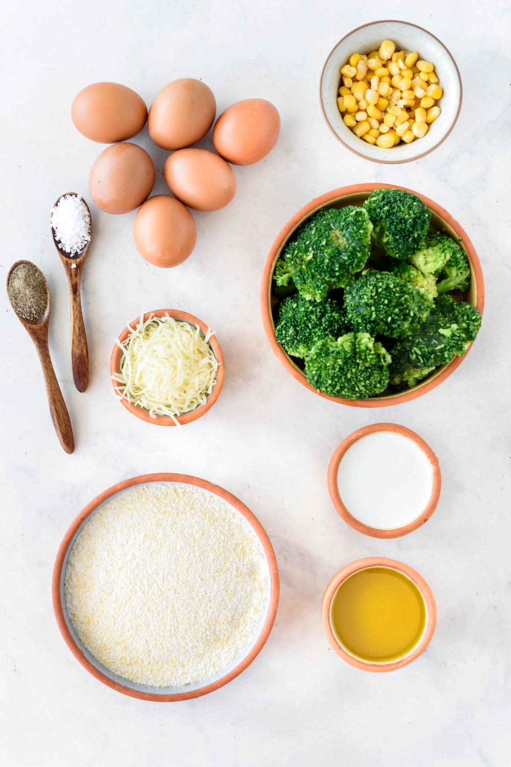 An assortment of ingredients including bowls of broccoli, corn, cornbread mix, milk, shredded cheese, and six whole eggs.