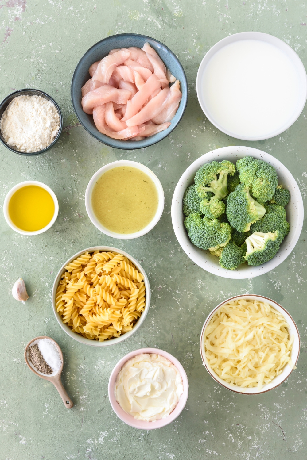 An assortment of ingredients including bowls of raw chicken strips, milk, shredded cheese, fresh broccoli, dried pasta, broth, oil, and spices.