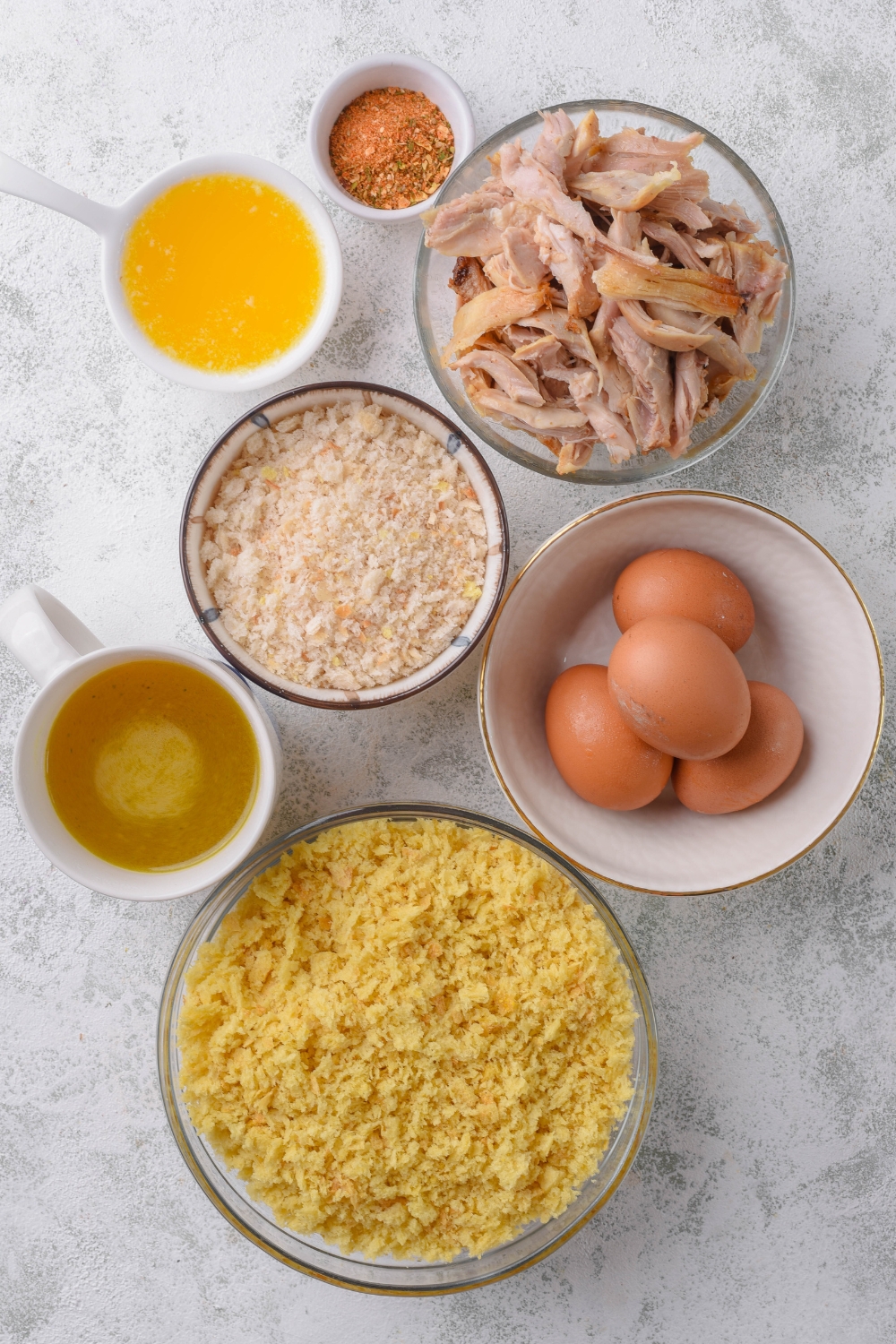 An assortment of ingredients including bowls of cornbread crumbles, four eggs, shredded chicken, bread crumbs, stock, spices, and melted butter.