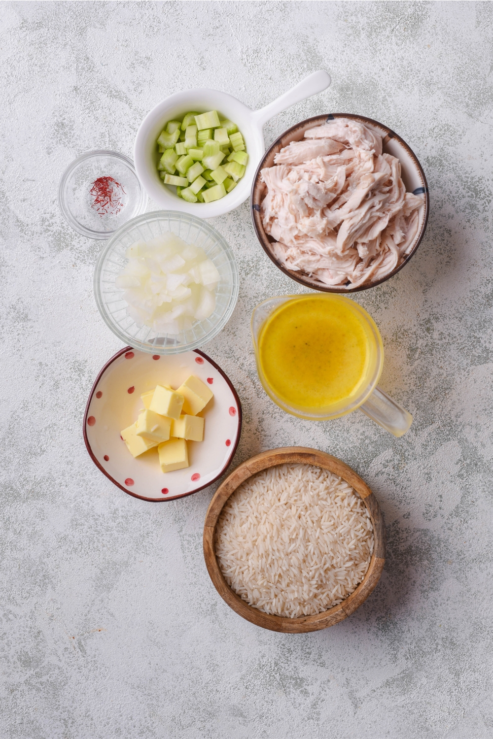 An assortment of ingredients including bowls of dried rice, broth, shredded chicken, butter cubes, diced onion, diced celery, and spices on a grey counter.